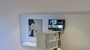 En grå soffa med två gula kuddar, en platt-TV som är monterad på väggen och en bokhylla under den.