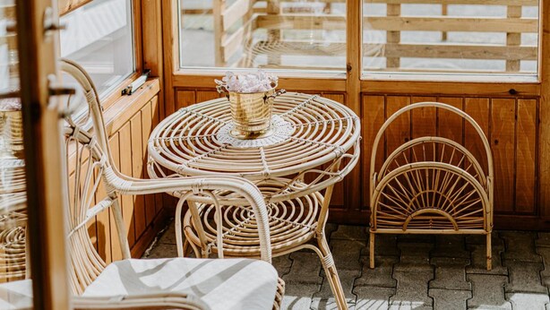 Una mesa de mimbre con sillas, un jarrón pequeño con flores y una valla de madera visible a través de la ventana.
