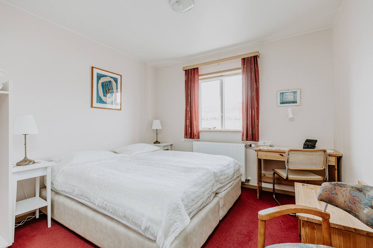 Hotellihuone, jossa on suuri sänky, työpöytä tuolilla, patteri ja ikkuna, jossa on verhot.
