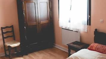 A bedroom with a bed, a wooden wardrobe, a chair, a small table, and a window with curtains.