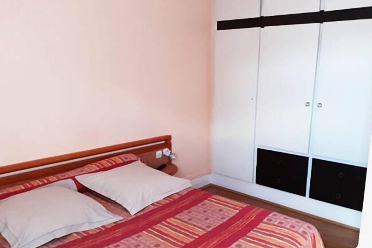 A bedroom with a bed, pillows, a wooden headboard, and a wardrobe with a black horizontal stripe.