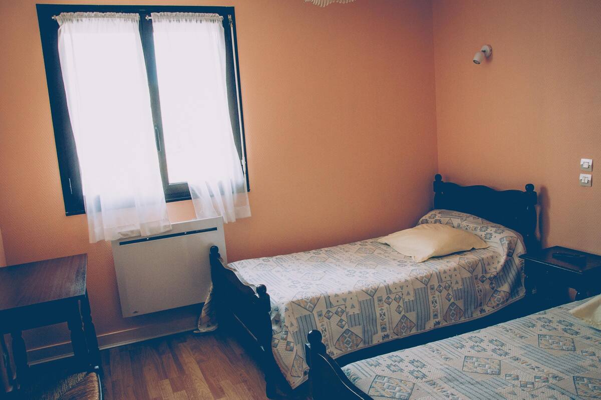 A small, single-bed room with a window, a radiator, a chair, and a bedside table.