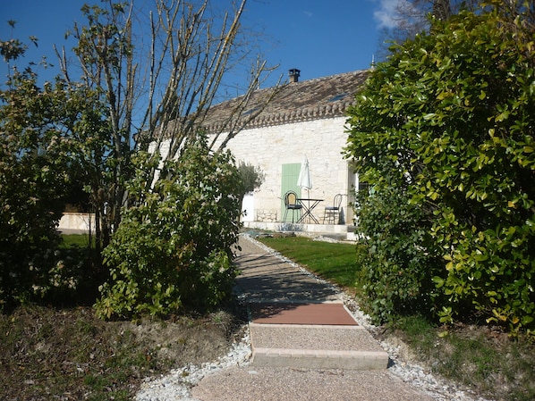 A stone building with a tiled roof, a paved pathway leading to a green door, and outdoor furniture.
