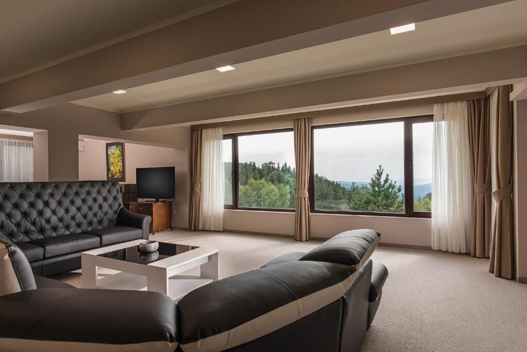 A modern living room with a black leather sofa, a white coffee table, and a television. Large windows offer a view of greenery and mountains.