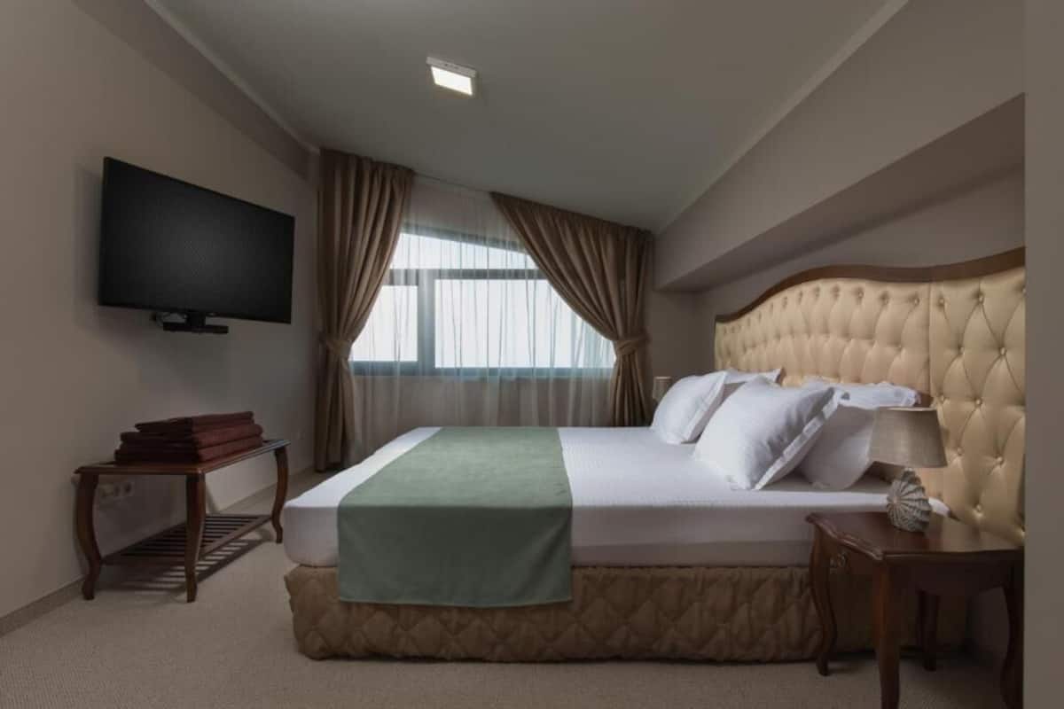 Una habitación de hotel con una cama grande, televisión, mesita de noche y ventana con cortinas.
