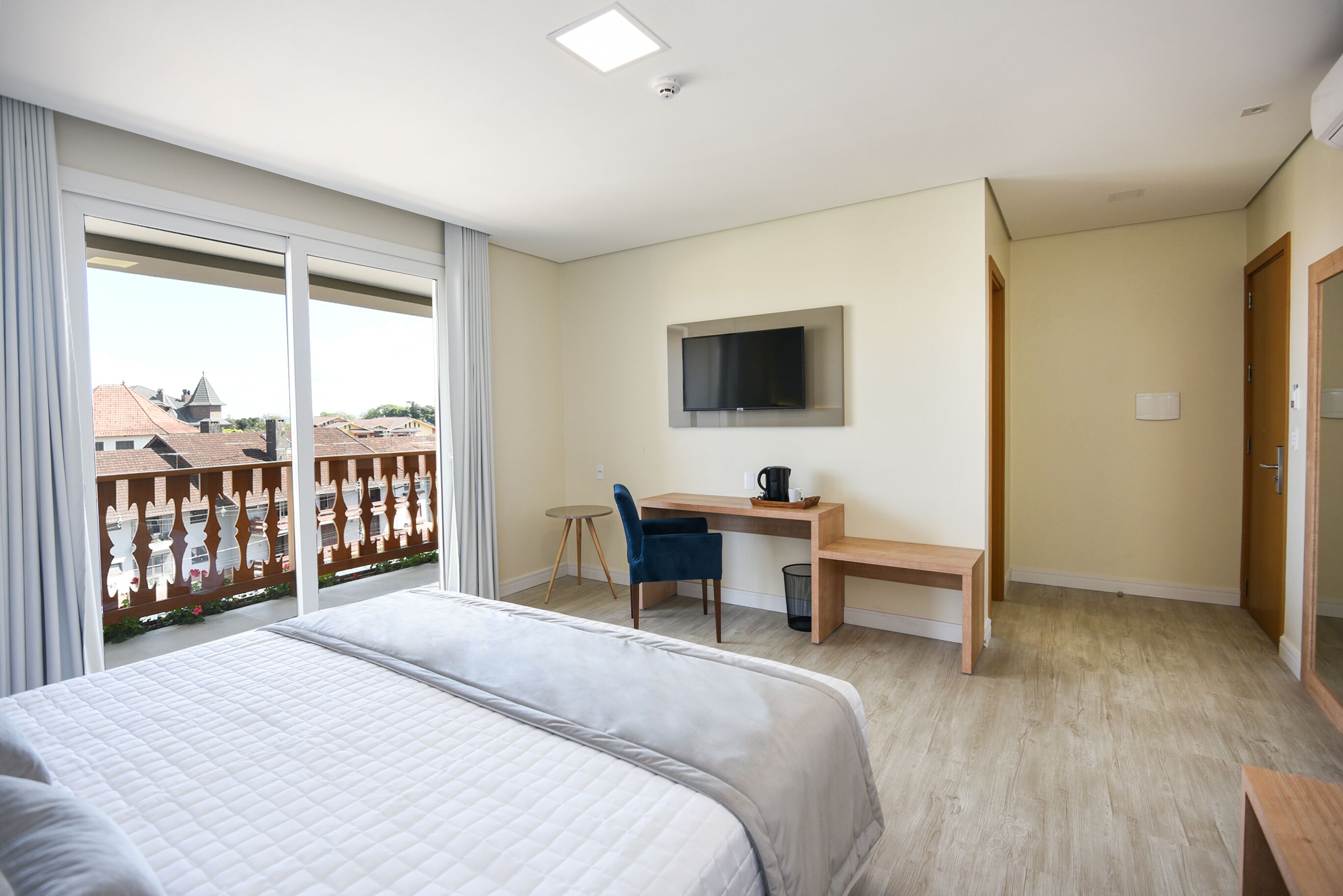 A hotel room with a bed, a desk, a chair, a TV, and a balcony with a view of buildings.