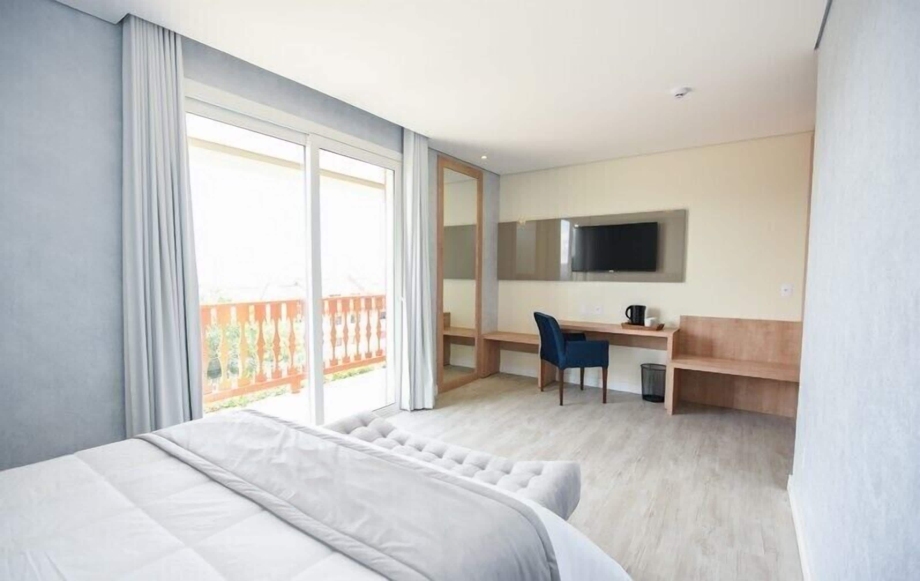 A hotel room with a large bed, a desk with a chair, a television mounted on the wall, and a balcony with a railing.