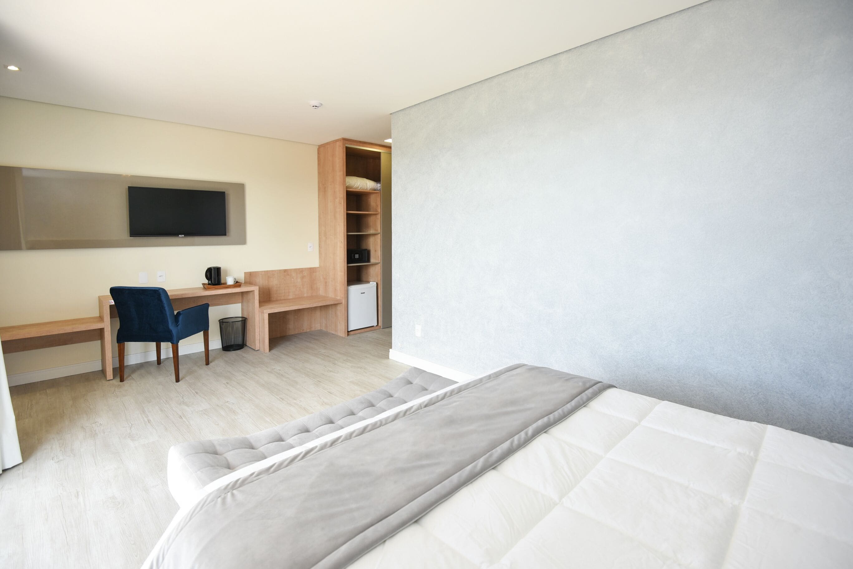 Quarto de hotel moderno com cama, escrivaninha, cadeira, televisão e armário embutido.