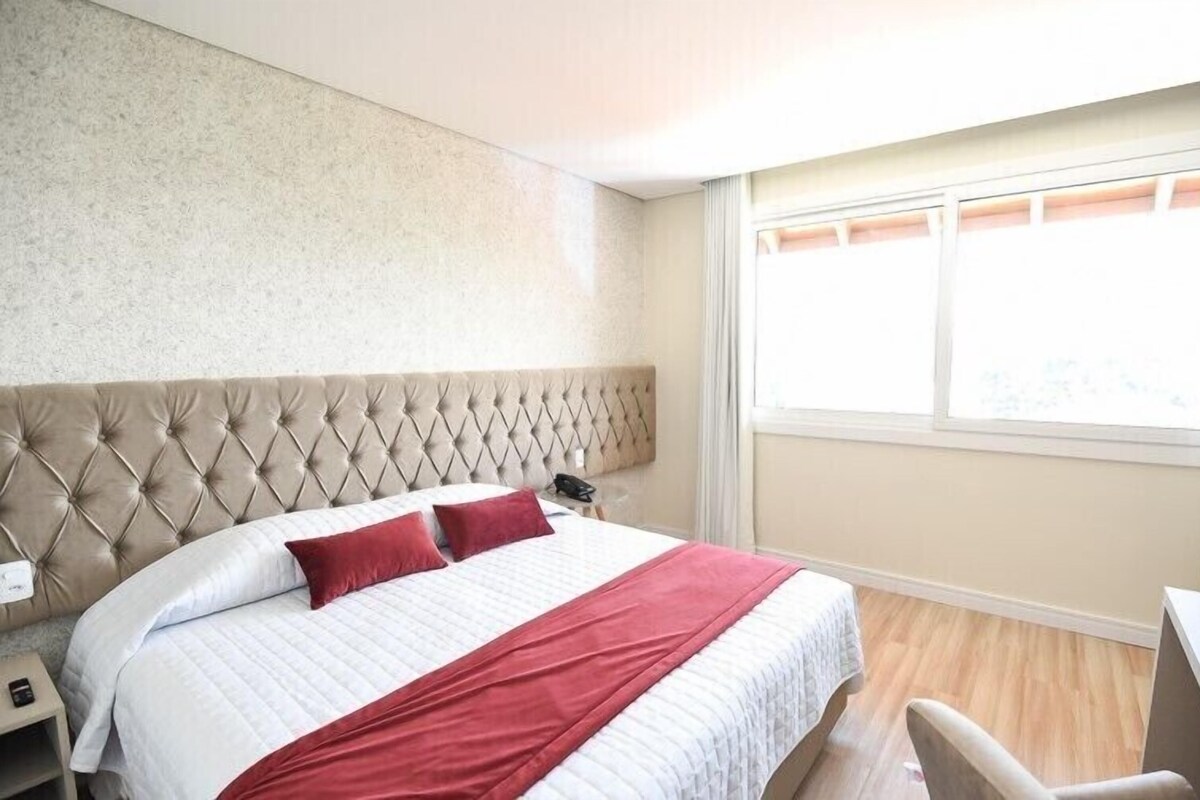 Una habitación de hotel con cabecera capitonada, una cama con ropa de cama blanca y roja, un escritorio y una ventana con persianas.
