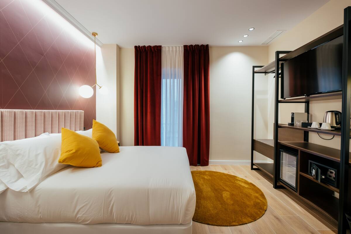 Habitación de hotel con cama, televisión, estante con objetos y ventana con cortinas.