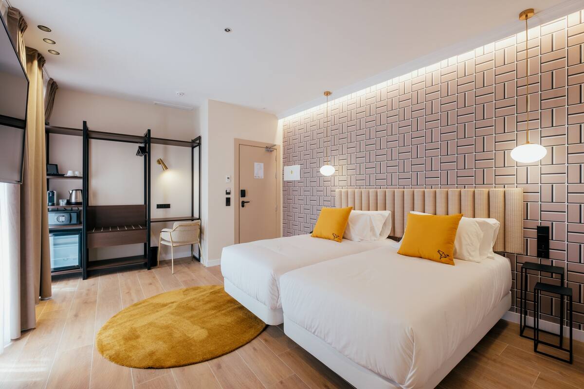 A hotel room with two beds, a wooden floor, a brick-patterned wall, and a small desk with a chair.