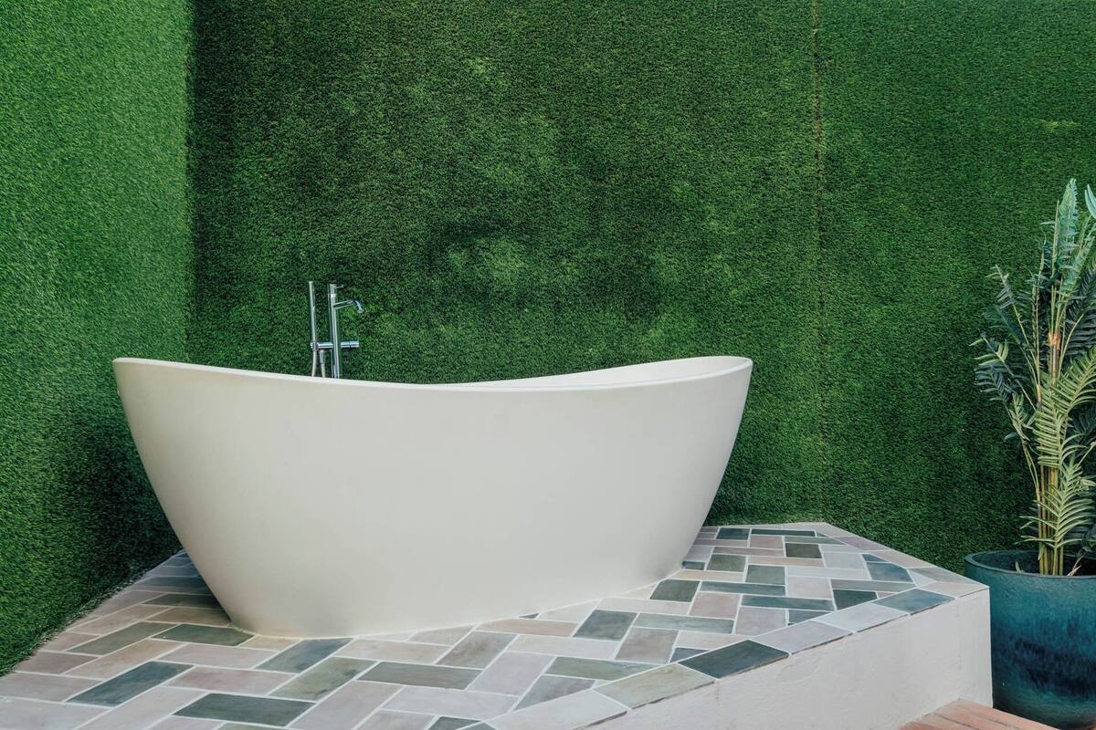 A freestanding bathtub with a modern design, positioned against a wall of artificial grass, on a tiled platform.