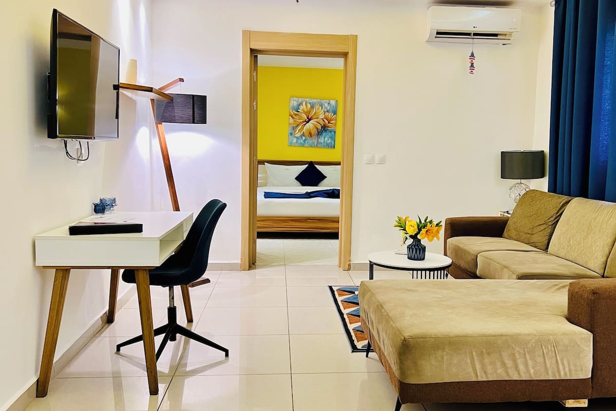 Una habitación de hotel moderna con escritorio, silla, sofá y cama, pared amarilla y un cuadro.