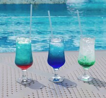 Three glasses of colorful drinks with straws, placed on a perforated metal table by a pool.