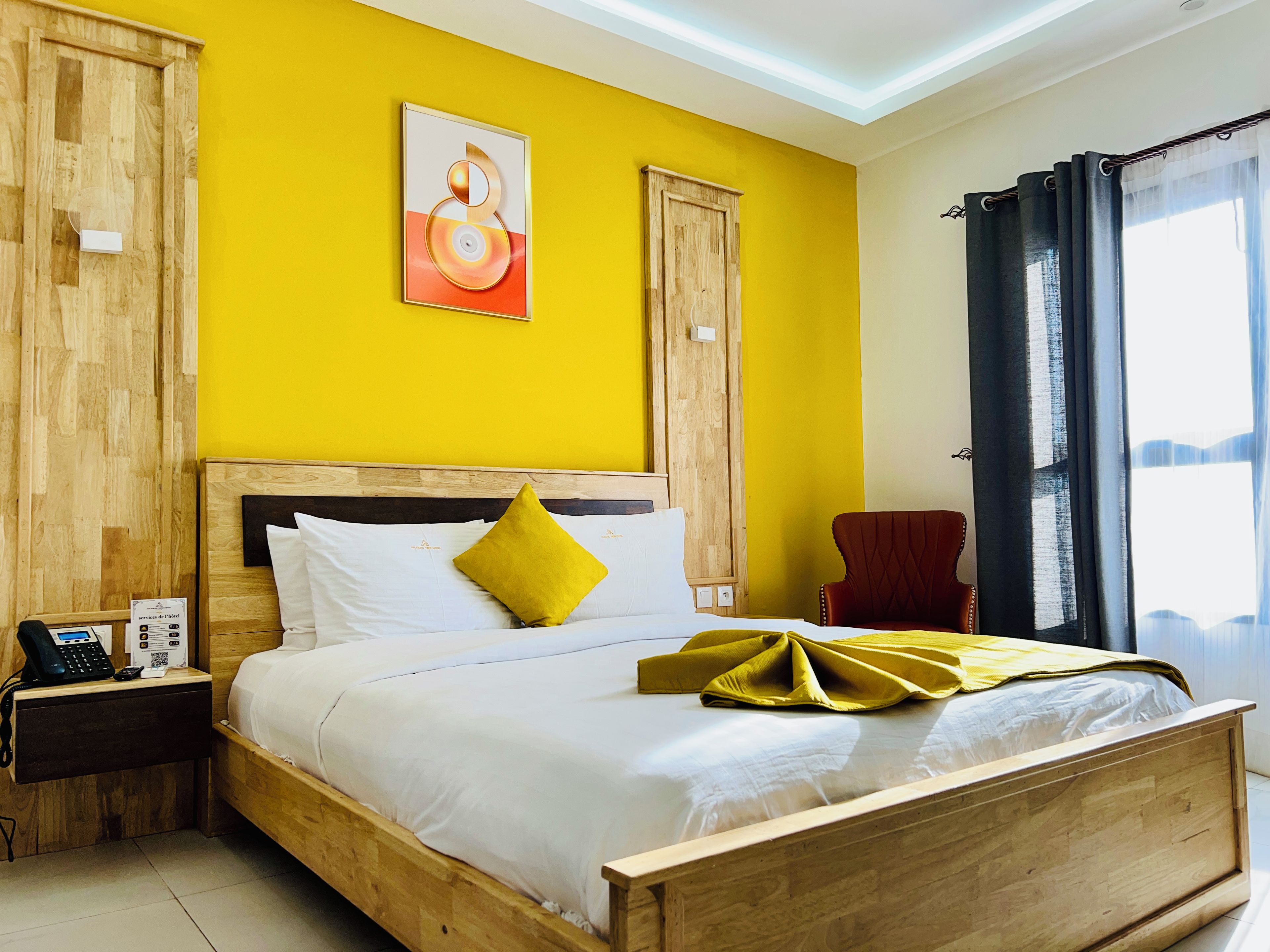 Habitación de hotel con cama de madera, mesita de noche con teléfono, una silla roja y ventana con cortinas.