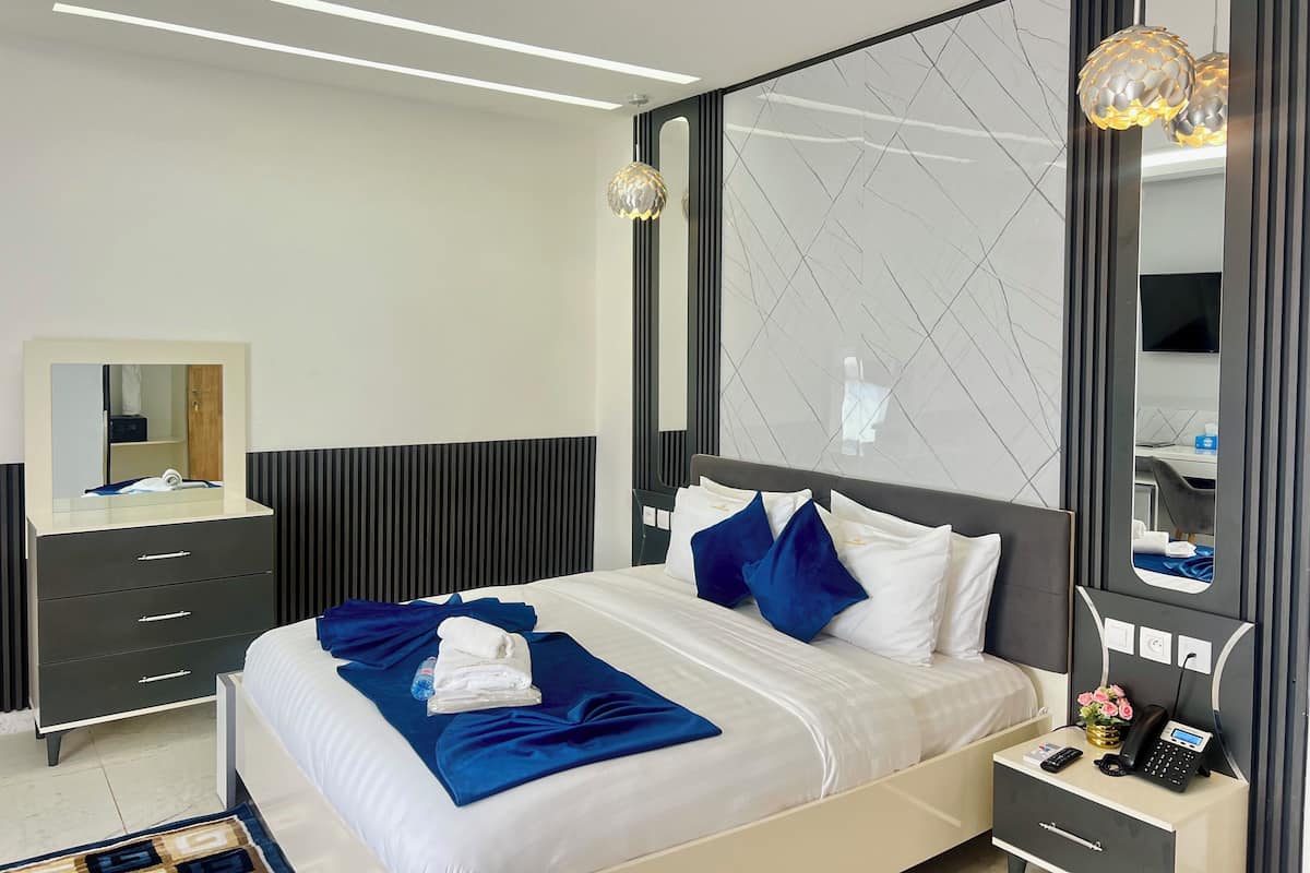 Habitación de hotel moderna con una cama bien hecha, una mesita de noche con teléfono y un jarrón de flores, un espejo grande y un televisor montado en la pared.