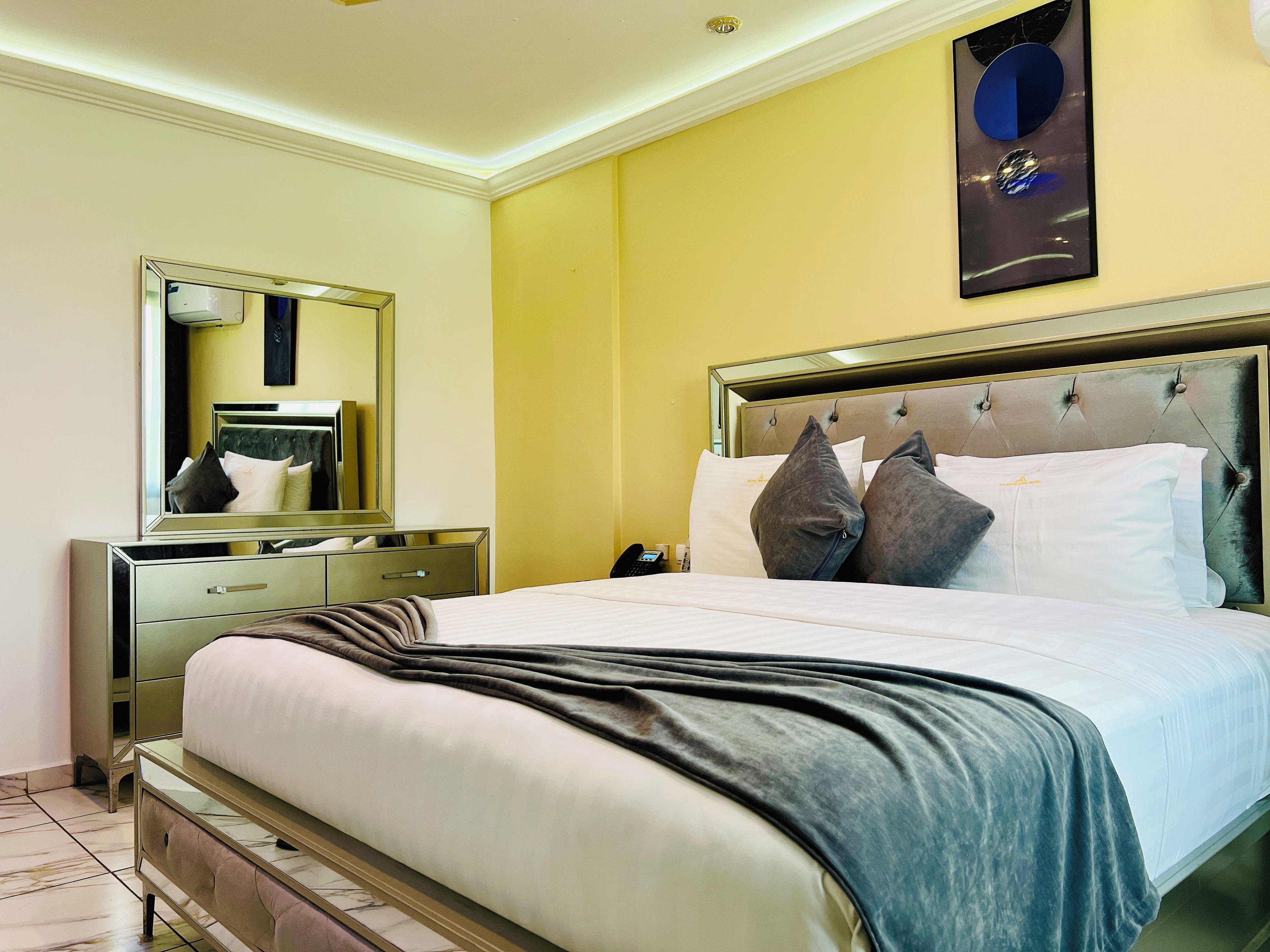 Quarto de hotel com uma cama grande, mesa de cabeceira, espelho e uma obra de arte na parede.