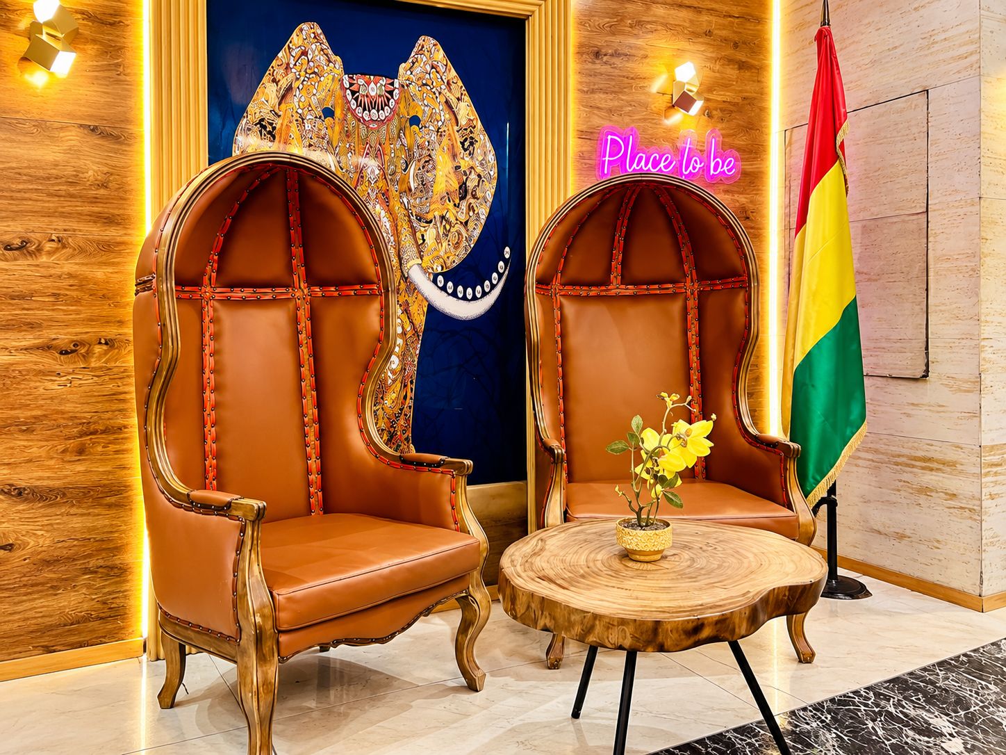 Two ornate leather armchairs with a round wooden table between them, a flag, and a neon sign in a room with wooden paneling and a tiled floor.