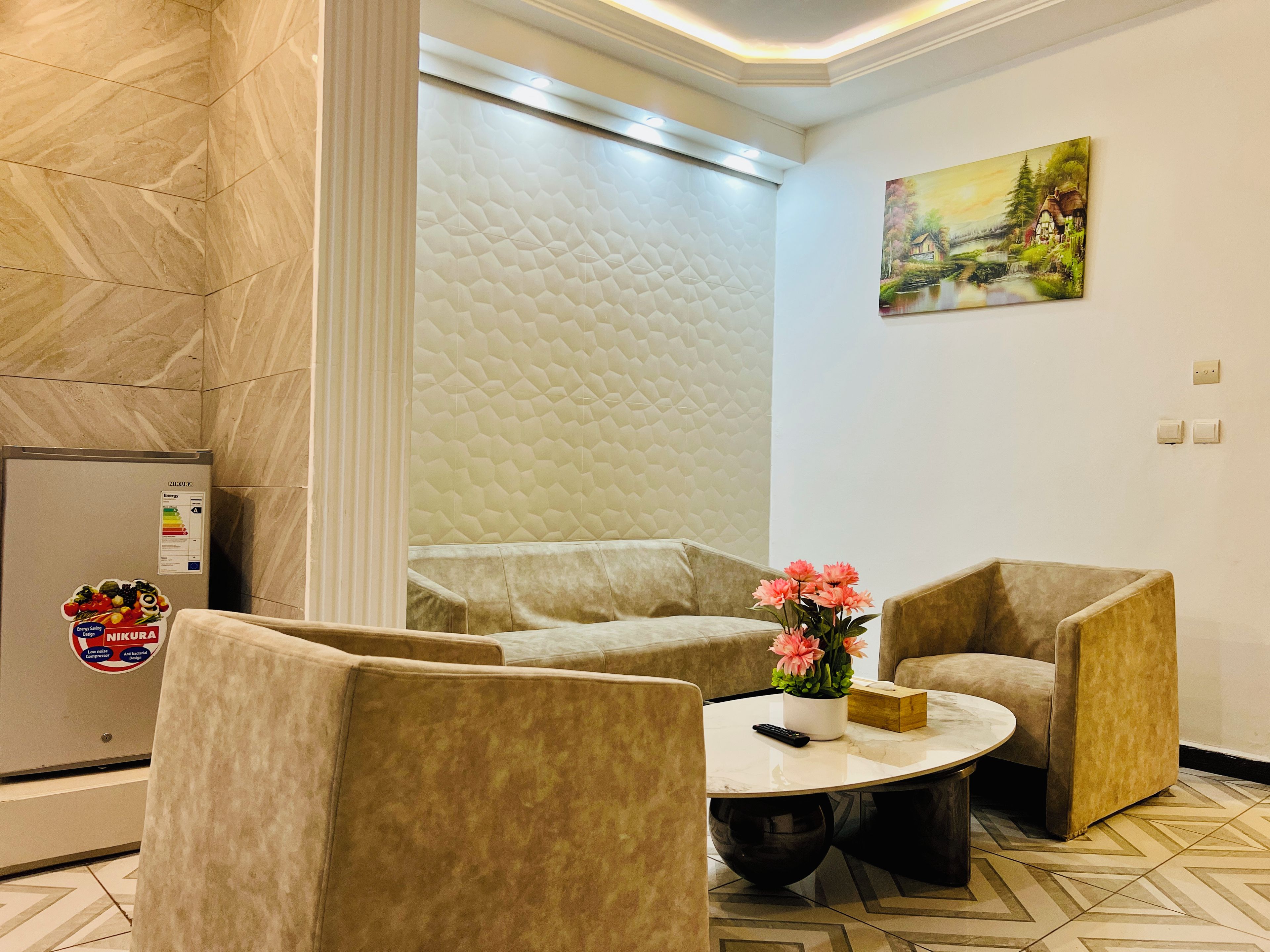A hotel room with a marble-patterned floor, a cream-colored sofa, a round coffee table with a vase of flowers, and a painting on the wall.