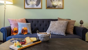 A dark blue sofa with multiple pillows, a small table with a basket labeled "PAIN," two glasses of rosé wine, cheese, and fruit. - A Florence Palace - 4 Bedroom Apt - Santa Croce (Florence)