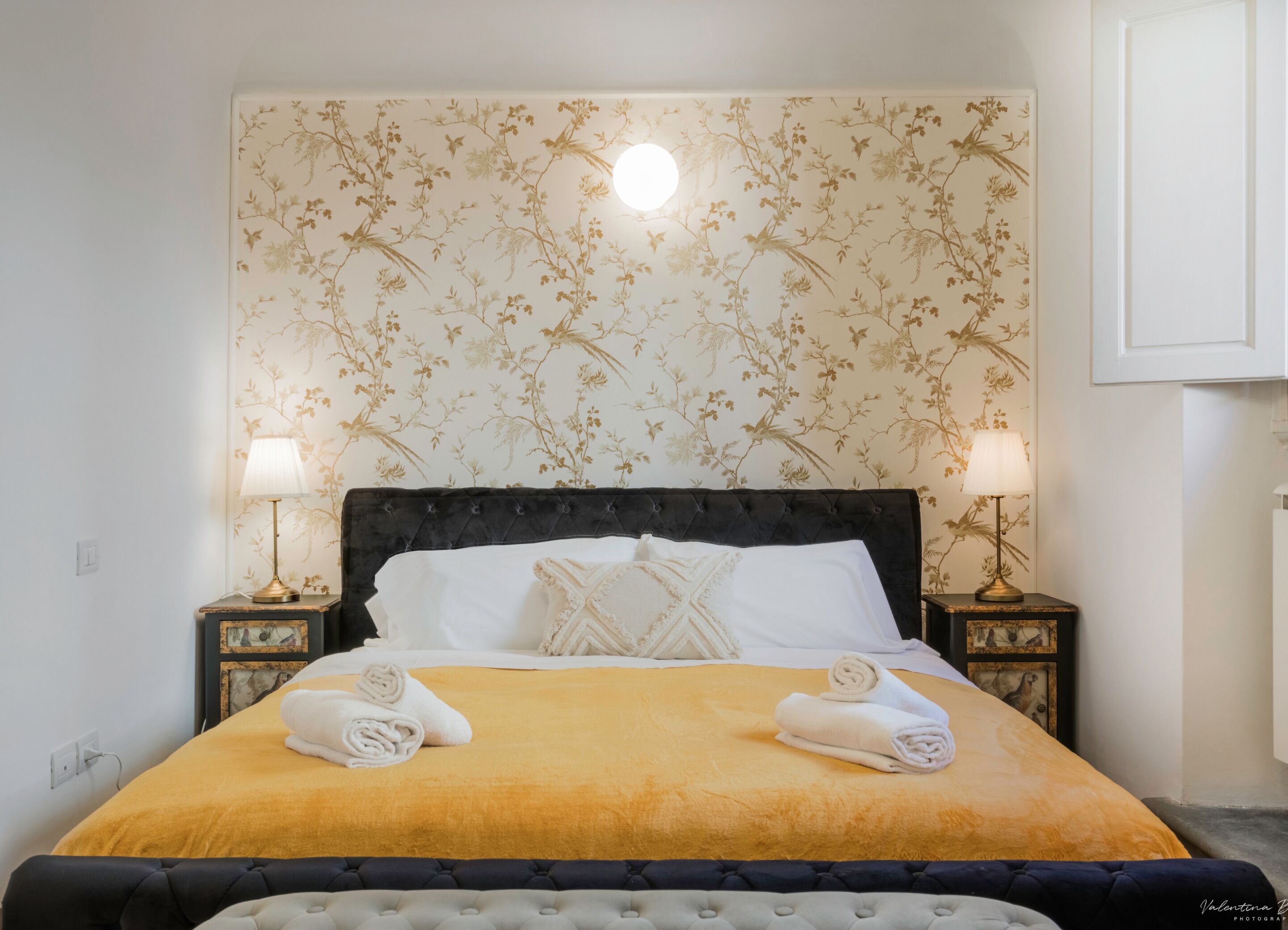 A neatly made bed with a yellow bedspread, two white pillows, and two rolled towels on the side. There are two bedside lamps on matching nightstands, one of which has a picture frame. The wall behind the bed features a floral wallpaper pattern.