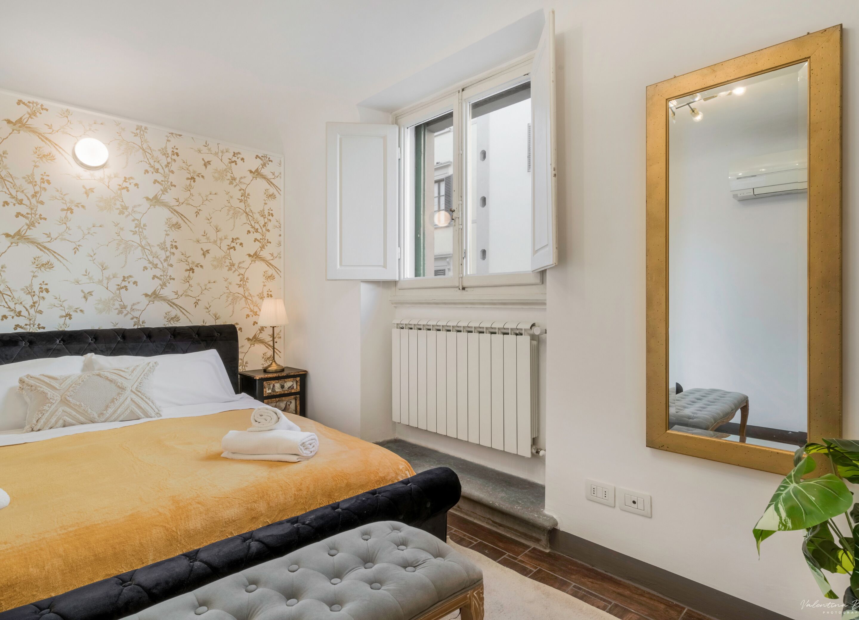 Een slaapkamer met een bed, een nachtkastje, een radiator, een raam met open luiken en een grote spiegel.