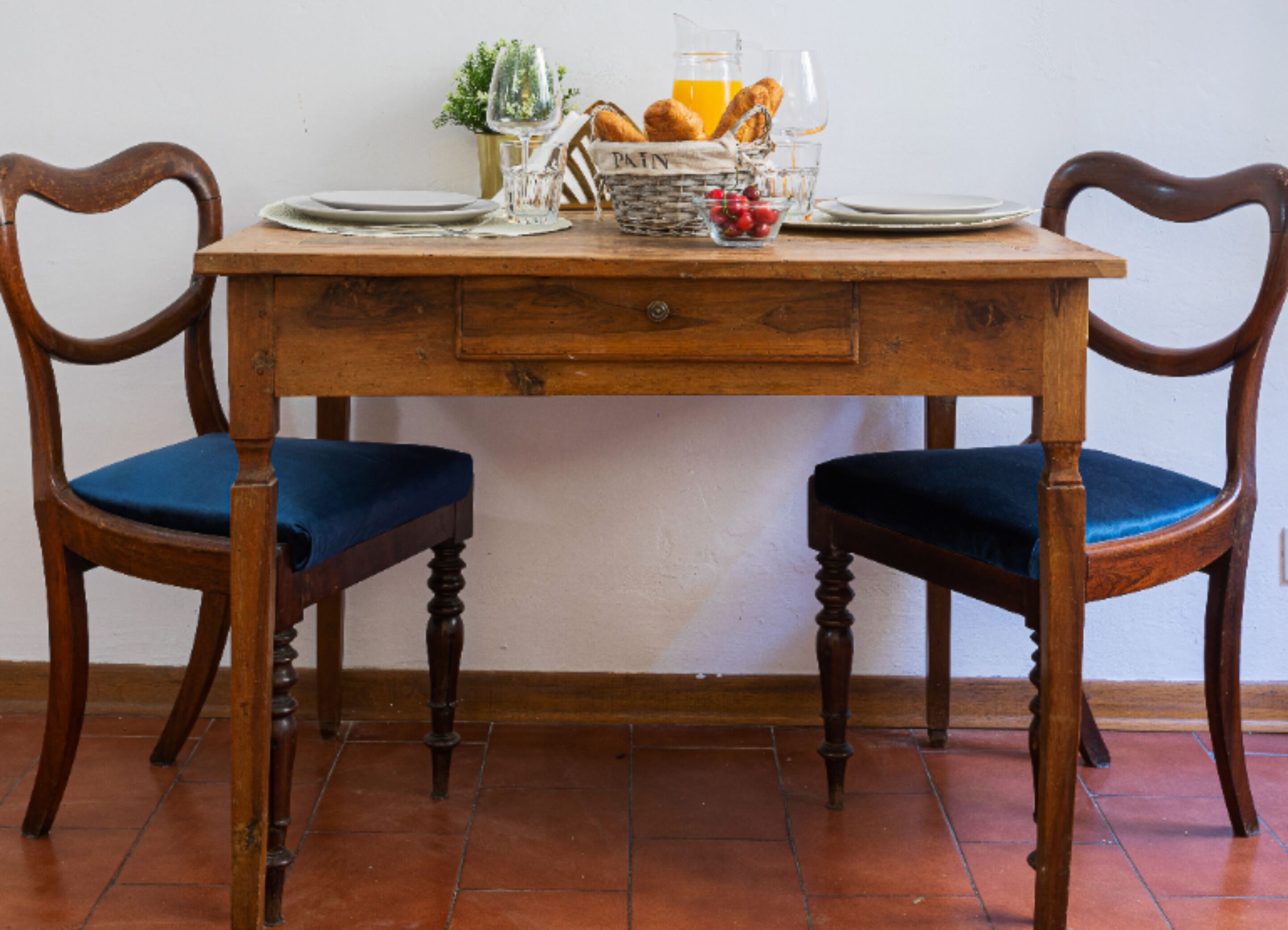 A wooden dining table with two chairs, a basket of bread, a glass of orange juice, and a pitcher.