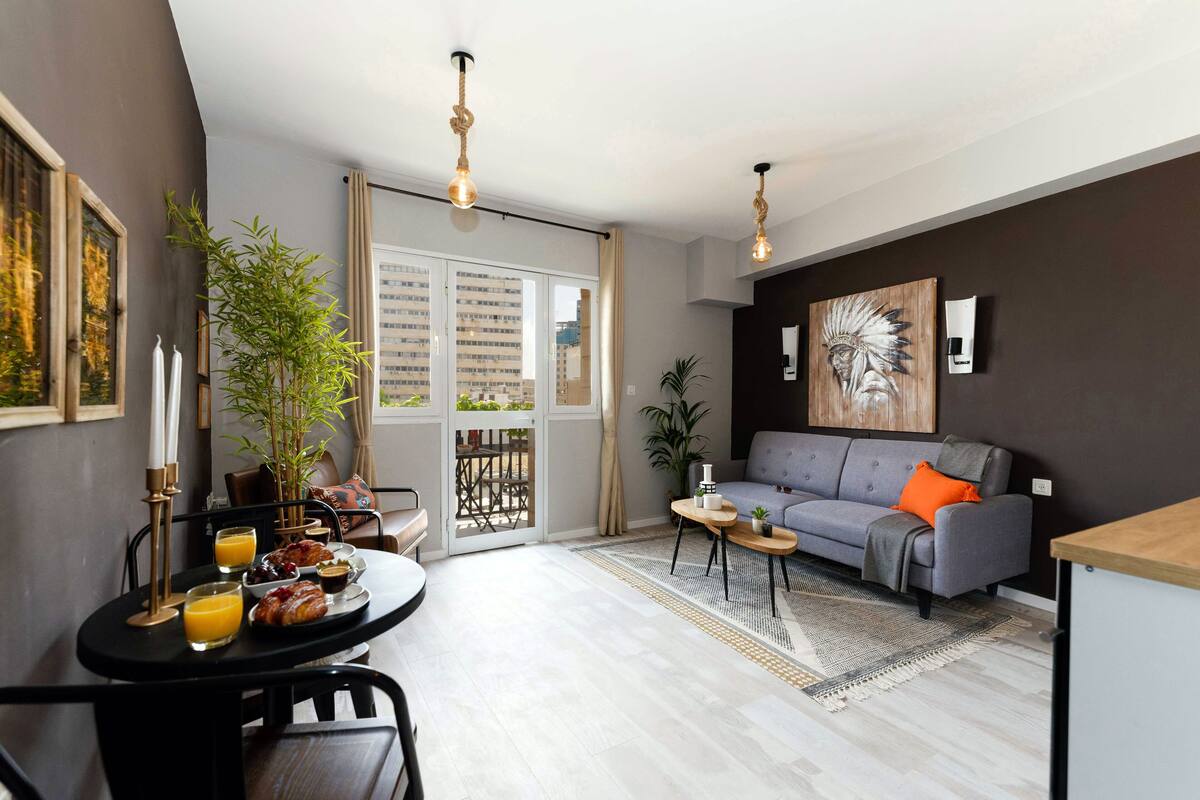 A modern living room with a grey sofa, a dining table with breakfast, and a large window.