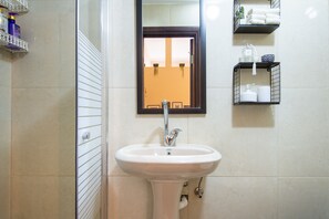 Casa de banho com lavatório branco, espelho e prateleiras com artigos de higiene pessoal.