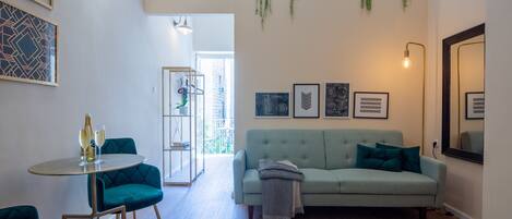 A modern living room with a green sofa, teal armchairs, and a round table with glasses. There are framed pictures on the wall and plants hanging from the ceiling.