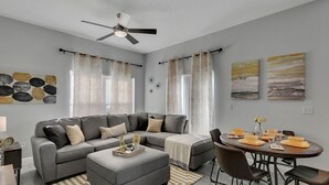 A modern living room with a gray sectional sofa, a dining table with chairs, and a ceiling fan. - 4 Bedroom Townhouse, Resort, 15 Mins to Disney, Themed Rooms perfect for Kids (Kissimmee)