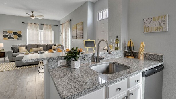 A modern kitchen with granite countertops, a stainless steel sink, and a dishwasher. There is a dining area with a gray sofa and a coffee table. - 4 Bedroom Townhouse, Resort, 15 Mins to Disney, Themed Rooms perfect for Kids (Kissimmee)