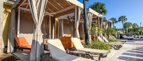 A poolside area with sun loungers and covered seating areas, palm trees, and a clear sky.