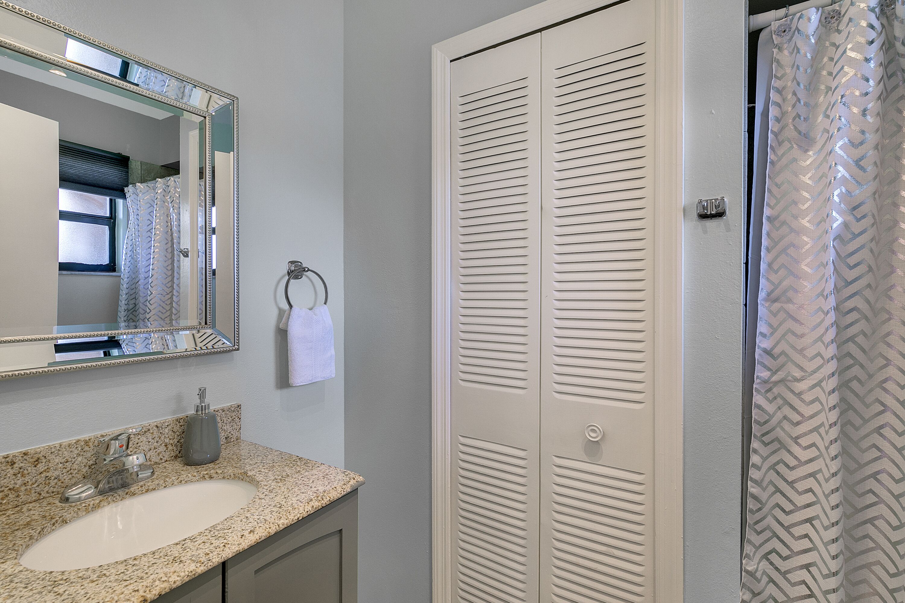 A bathroom with a granite countertop sink, a mirror, a towel rack, and a white closet with louvered doors.