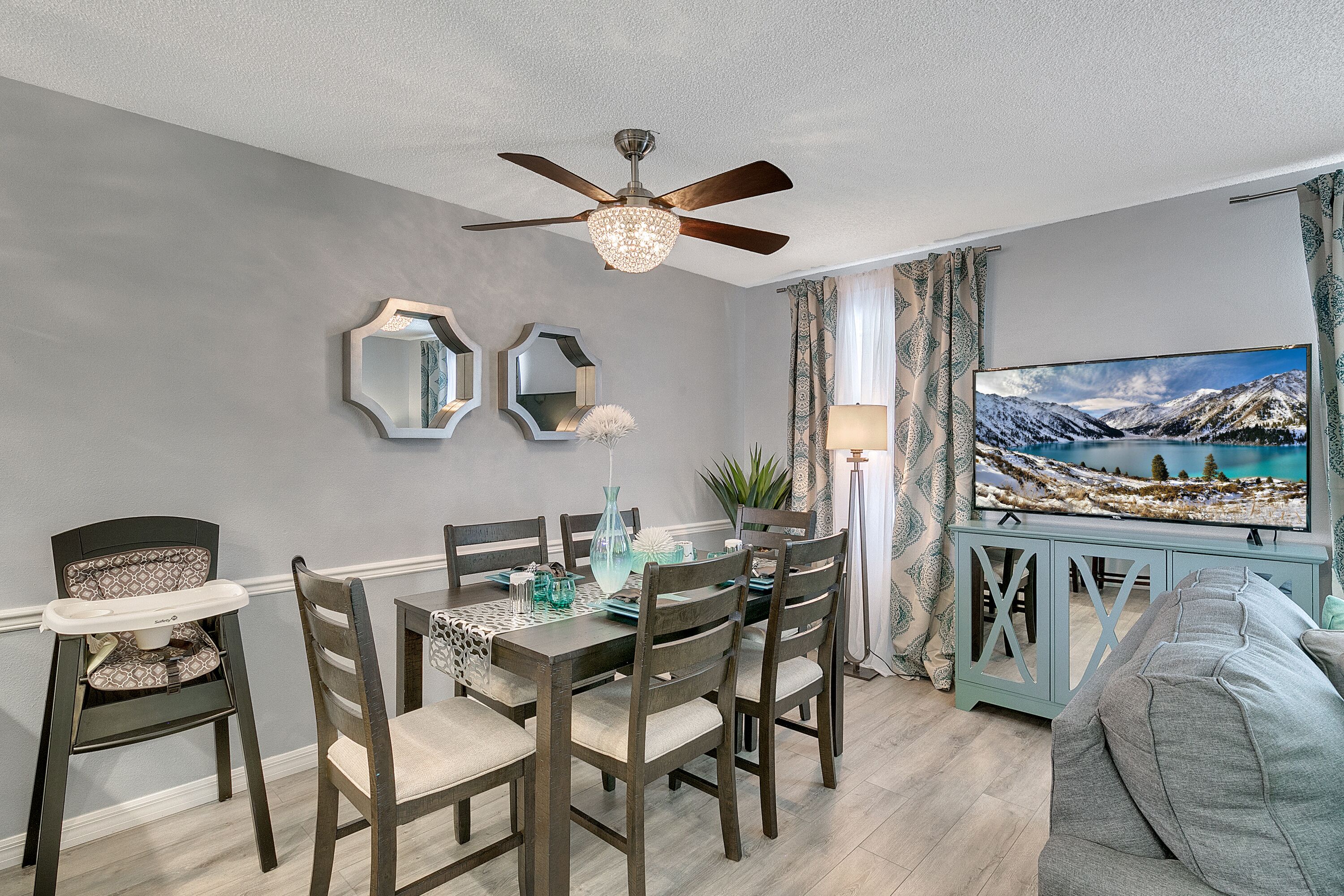 Un coin repas comprenant une table, des chaises, un ventilateur de plafond et un téléviseur diffusant un paysage montagneux.