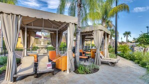 A covered outdoor seating area with lounge chairs and a table, surrounded by palm trees and a fountain. - Family Vacation Home, Water Park+Endless Amenities (Kissimmee)