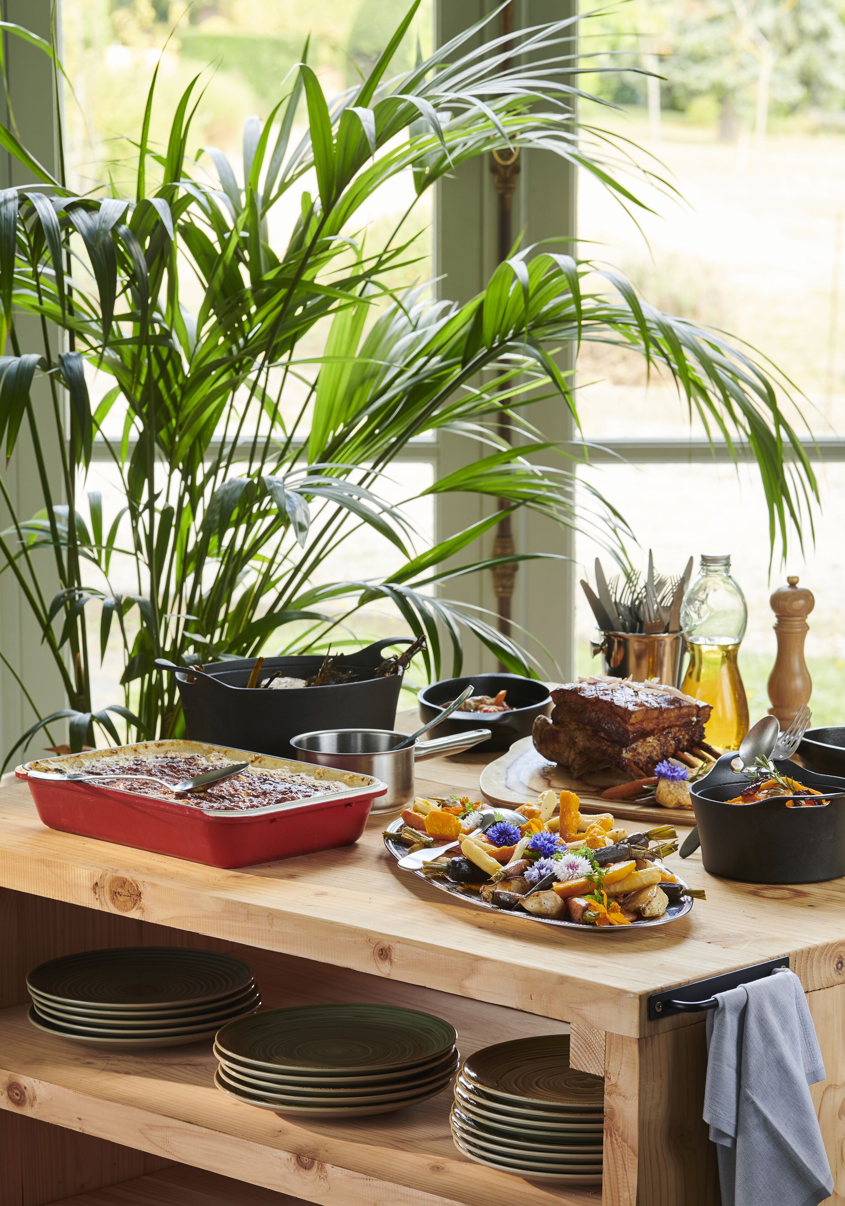 A wooden table with a variety of dishes, a red baking dish, and a large plant.