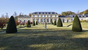 Un grand bâtiment de style classique, doté d’une architecture symétrique, d’une pelouse bien entretenue et d’arbres taillés en formes géométriques.