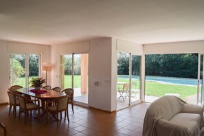 A spacious living area with a dining table and chairs, a covered sofa, and large sliding glass doors leading to a garden and pool. - Villa 100 meters from the sea, in the Platja de Pals Golf Club. A/C (Pals)