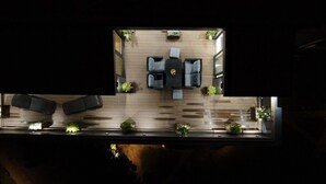 Vista cenital de una zona de estar al aire libre moderna, con terraza de madera, mobiliario exterior negro y plantas en macetas.
