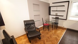 A room with a wooden herringbone floor, a black leather chair, a glass table with two chairs, and a radiator. Shelves with decorative items are mounted on the wall. - Metropol (Belgrade)