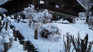 Um chalé de madeira com telhado inclinado, rodeado por árvores cobertas de neve e um caminho nevado que leva à entrada.