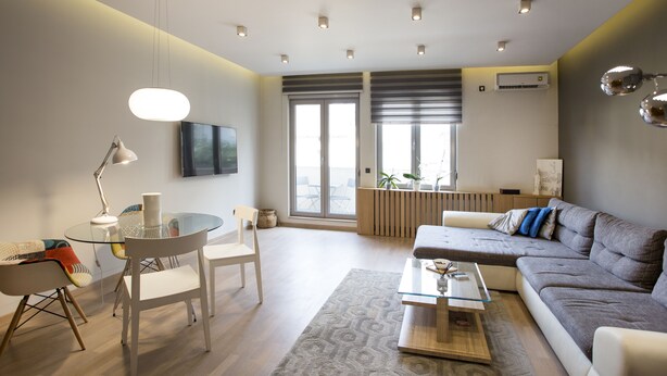 A modern living room with a glass dining table, a sofa, and a coffee table. There is a TV mounted on the wall and a window with blinds.
