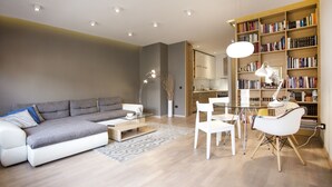 A modern living room with a grey sofa, white chairs, a glass coffee table, a bookshelf, and a dining area with a table and chairs.