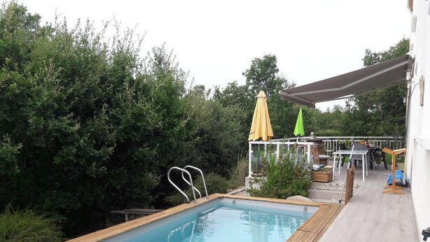 An outdoor swimming pool with a wooden deck, surrounded by greenery. - La Maison Familiale (Lagarrigue)