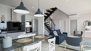 A modern kitchen with a central island, white chairs, and pendant lighting. A staircase with wooden railings leads to a living area with a blue sofa and a round mirror. - Felicidade Rocha II - For families, with a pool (Ponta do Pargo)