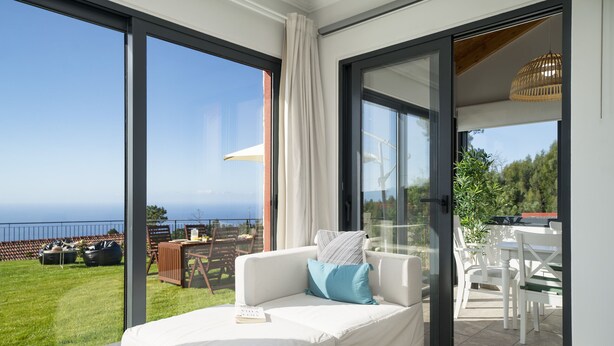 A modern living room with a white sofa, a book, and a view of a grassy area and the sea. - Felicidade Rocha II - For families, with a pool (Ponta do Pargo)