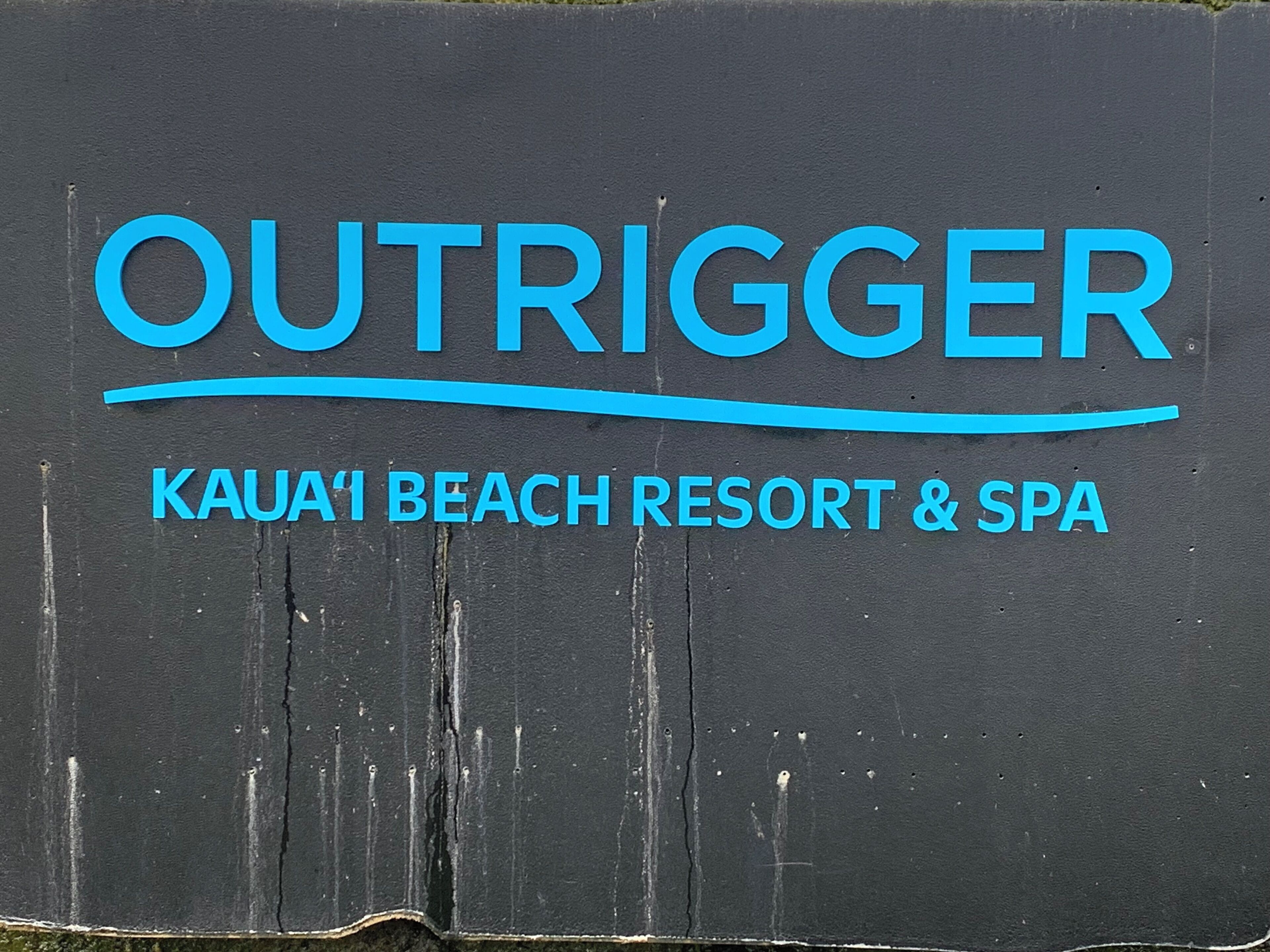 A sign for OUTFITTER KAUAI BEACH RESORT & SPA with a wooden background and blue lettering.