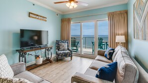 A living room with a TV, a ceiling fan, a sofa, and a view of the ocean through large sliding doors. - Gulf Shores condo with fabulous views of the Gulf of America (Gulf Shores)