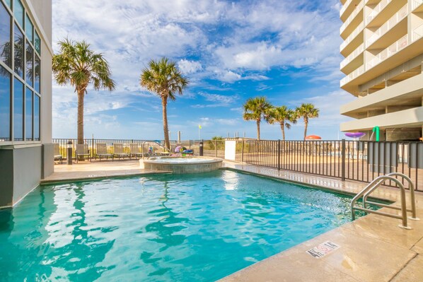 A swimming pool with a hot tub, surrounded by palm trees and a fence, with a clear sky in the background. - Gulf Shores condo with fabulous views of the Gulf of America (Gulf Shores)